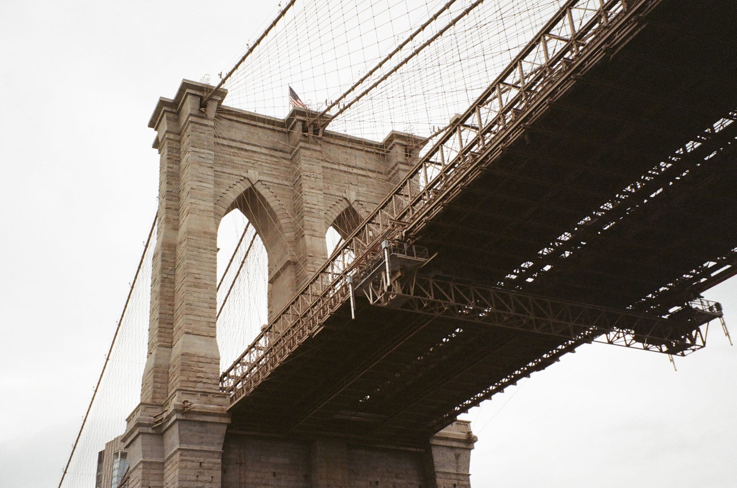 Looking up at a stone tower and the underside of the Brooklyn Bridge under an overcast sky