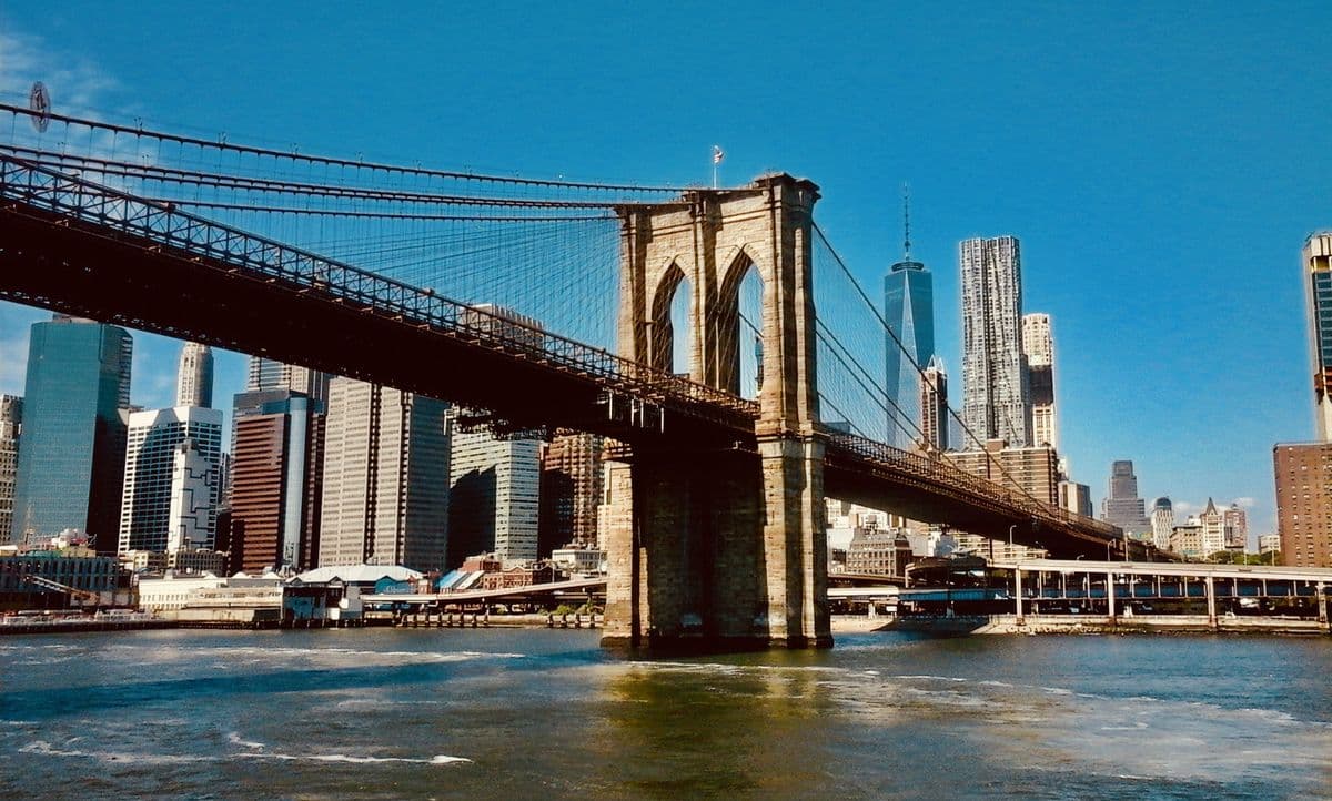 The Brooklyn Bridge and its stone tower under a clear blue sky with the Manhattan skyline behind
